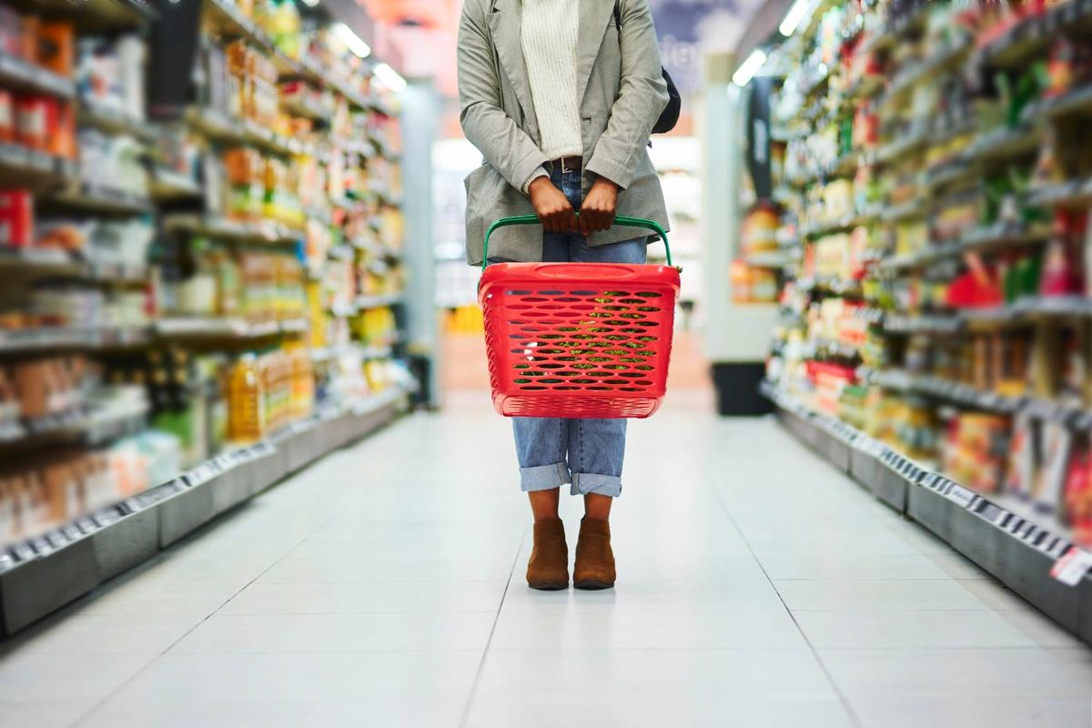 A stock image of a woman standing in the supermarket, holding an empty red shopping basket.