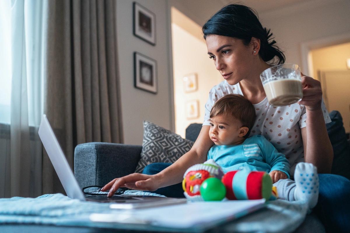 A single mother works on a laptop with her young son sat on her knee.