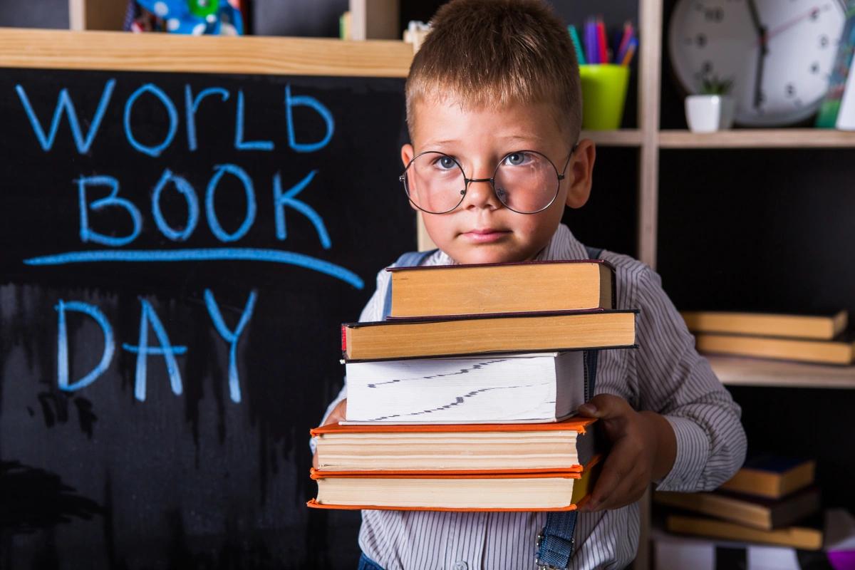 A stock image of a little boy with a stack of books in front of a chalkboard with 'WORLD BOOK DAY' written on it in blue chalk.