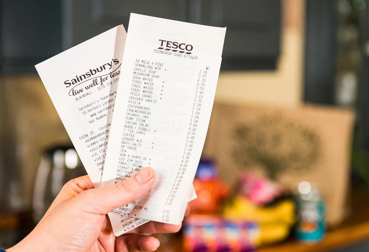 A stock image of a hand holding receipts from Sainsbury's and Tesco in front of blurred out groceries.